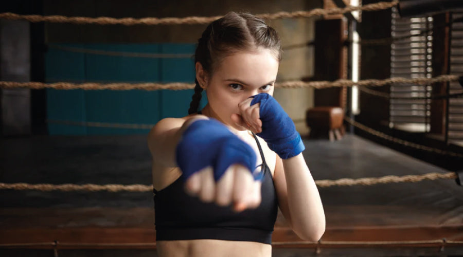 Young girl facing with braids and wearing a black sports bra is standing in front of a dark boxing ring with blue gloves on punching with her right hand towards the camera